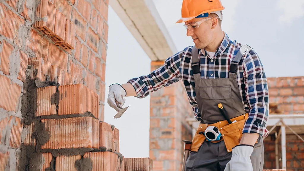 Un homme en salopette et casque travaille sur des briques pour construire une maison.